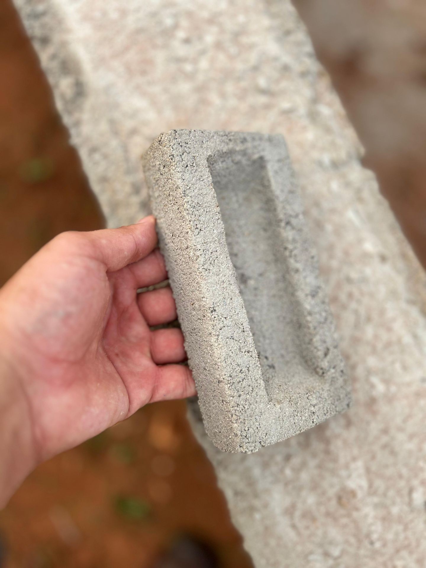 Hand holding a small rectangular cement block with a hollow center, against a blurred background.